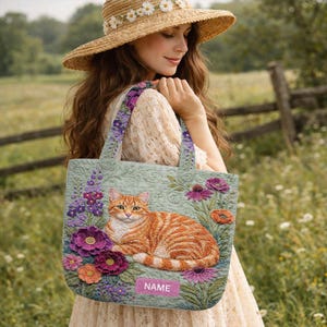 May include: A light green tote bag featuring an orange tabby cat surrounded by embroidered flowers in shades of purple, pink, and orange. The bag has purple floral straps and a small pink label with the word "NAME".