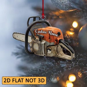 May include: A flat, two-dimensional chainsaw ornament featuring an orange and white body with a black handle and a silver blade. The side of the chainsaw displays the text Custom Name in a white script font, and it hangs from a thin red string.