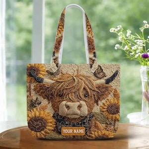 May include: A beige tote bag featuring a Highland cow design with sunflowers and butterflies. The bag has a white handle and the text "YOUR NAME" on a small rectangular tag.