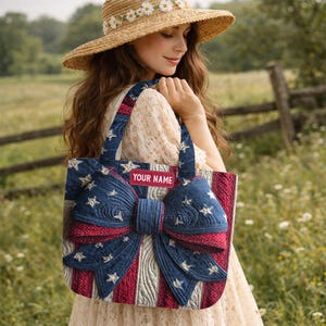 May include: A patriotic tote bag with a large bow design. The bag features a blue background with white stars and red and white stripes. The text "YOUR NAME" is displayed on a red label. The bag has two handles.