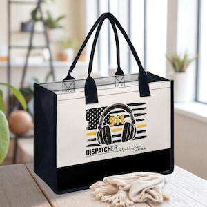 May include: A tote bag with black handles and trim, featuring a design for a 911 dispatcher. The design includes a stylized American flag, headphones, the number 911, and the word "DISPATCHER". The bag is on a wooden surface.