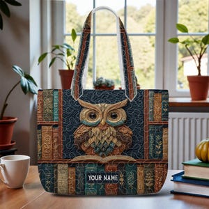 May include: A quilted tote bag featuring a detailed owl design with an open book, set against a backdrop of bookshelves. The bag has a navy blue background, with the text "YOUR NAME" on the book. The handles are white with brown and blue accents.