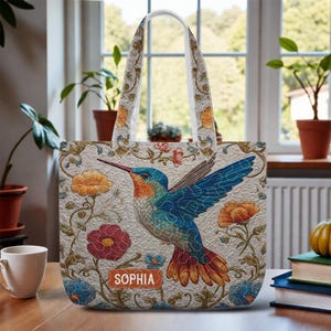 May include: Beige tote bag featuring a colorful hummingbird design with blue wings, a long beak, and floral accents. The bag has a white handle and the name "Sophia" is printed on the front.