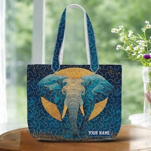 May include: A tote bag featuring an elephant design. The elephant is depicted with blue and gold accents against a navy blue background with a quilted pattern. The bag has a white handle with blue trim and the text "YOUR NAME".