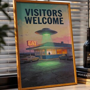 May include: A framed poster with the words "VISITORS WELCOME" above an illustration of a diner being illuminated by a UFO. A bottle of wine and a stack of books are on a windowsill next to the poster. The poster has a retro, vintage aesthetic.