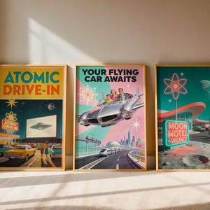 Three framed retro-futurist posters in wooden frames against a beige wall. Left: yellow and teal 1950s drive-in theater with UFO on screen. Center: pink and teal family in a chrome flying car above a futuristic cityscape. Right: teal lunar motel with neon vacancy sign, rockets, and Earth in the background.