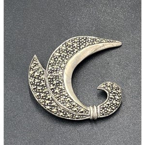 May include: A silver-toned marcasite brooch in a stylized, curved design. The brooch features a crescent shape with a textured surface of small, sparkling stones. The design includes a leaf-like element and a curled end.