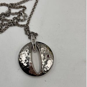 May include: A silver-toned necklace featuring a circular pendant with a hammered, textured finish. The pendant has a central vertical cutout and a small, square-set red gemstone on the right side. It hangs from a matching silver-toned link chain.