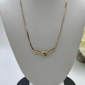 May include: A gold-toned necklace featuring a thin, flat chain and a central V-shaped pendant. The pendant is accented with a small, iridescent white oval stone and a tiny, dark red circular stone, creating a subtle, refined focal point.