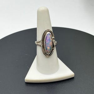 May include: A silver ring featuring an elongated oval-shaped iridescent opal gemstone set in a decorative metal bezel. The band is split at the shoulders and the setting is accented with small etched lines around the perimeter of the stone.