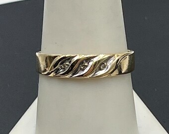 May include: A gold band ring featuring a central diagonal wave pattern in white metal. The design includes three small, round, clear stones set within the silver-toned diagonal grooves, contrasting against the polished yellow gold finish of the band.