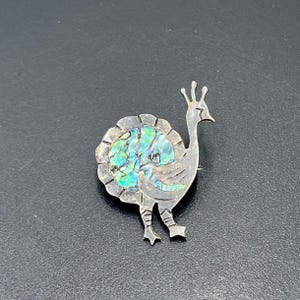May include: A silver-toned metal brooch shaped like a stylized bird, featuring a circular body inlay of iridescent abalone shell with vibrant green and blue hues. The bird has a tall crest, a pointed beak, and detailed etched lines on its legs.