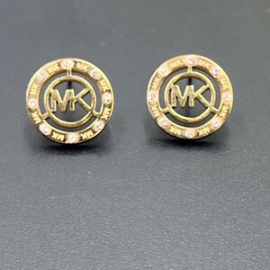 May include: A pair of gold-tone circular stud earrings featuring the letters MK in the center. The outer rim of each earring is decorated with small clear stones and the repeating text MK. The design is open-work with a polished metallic finish.