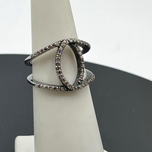 May include: A dark-toned metal ring featuring a central vertical oval shape. The band splits into two distinct sections that meet at the oval, with the entire surface of the oval and the split bands encrusted with small, clear, sparkling stones.