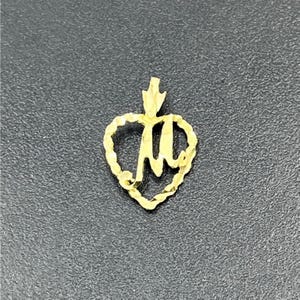 May include: A gold-colored pendant featuring a heart-shaped outline with a textured, scalloped edge. Inside the heart is a stylized, cursive letter M. The pendant includes a small, triangular bail at the top for attachment to a chain or necklace.