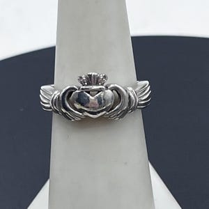 May include: A silver Claddagh ring featuring a central heart held by two hands, topped with a crown. The metal has a polished, reflective finish, showcasing the detailed design of the hands, the heart, and the crown elements on the band.