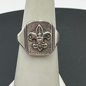 May include: A silver-toned ring featuring a rectangular face with a textured, hammered background. Centered on the face is a raised fleur-de-lis emblem with a shield detail. The band has a smooth, polished finish with vertical grooved accents.