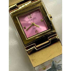 May include: A gold-toned Joan Rivers Classics quartz watch with a pink square face. The watch face displays the numbers 12 and 6, and the word "QUARTZ". The watch band has a floral design.