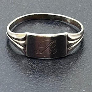 May include: A silver-toned metal ring featuring a rectangular face with a script letter R engraved in the center. The band has a split design on both sides, connecting to the central rectangular plate with a smooth, polished metallic finish.