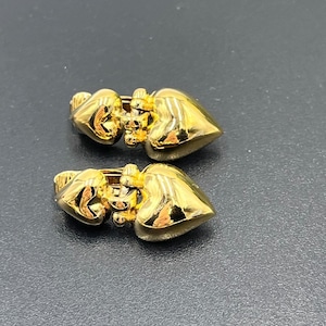 May include: A pair of gold-colored heart-shaped earrings. The earrings have a polished, reflective surface and a hinged clasp. The hearts are smooth and rounded, with a slight indentation at the top.