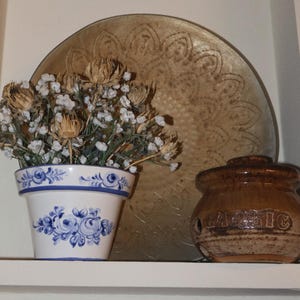 May include: A still life arrangement featuring a white and blue floral pot with dried flowers, a decorative gold-toned plate, and a brown ceramic garlic jar. The jar has the word "GARLIC" etched on its side.