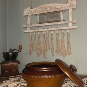 May include: A distressed white wooden wall hanging with a weaving loom and hanging tassels. A brown ceramic pot with a lid rests on a lace doily. A vintage coffee grinder is on the left.