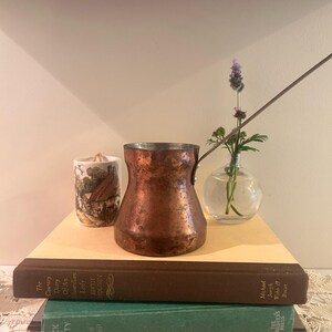 May include: A weathered copper coffee pot with a long metal handle sits atop a stack of two hardback books. Beside the pot is a small ceramic candle holder with a landscape illustration and a clear glass vase containing a single purple flower.