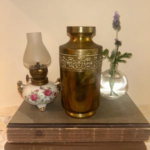 May include: A brass vase with a decorative embossed band around the centre, positioned next to a small porcelain oil lamp featuring pink floral patterns and a clear glass chimney, and a small clear glass bud vase containing a sprig of lavender.