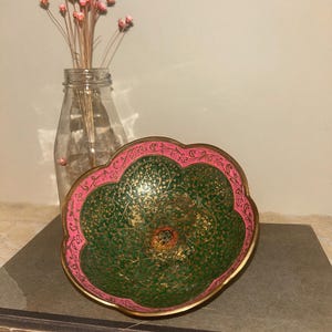 May include: A scalloped metal bowl featuring a vibrant green interior with intricate gold floral engravings and a bright pink border. The bowl is positioned in front of a clear glass bottle containing a small bouquet of dried pink flowers.