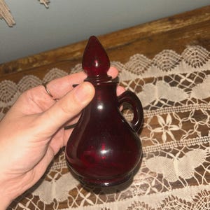 May include: A small, ruby-red glass bottle with a handle and a pointed stopper. The bottle has a rounded body and a small handle. The stopper is also red and has a teardrop shape. The bottle is likely vintage and decorative.