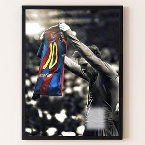 Puede incluir: Fotografía en blanco y negro enmarcada que muestra a un jugador de fútbol sosteniendo una camiseta. La camiseta es roja, azul y amarilla con el número 10. La imagen tiene un fondo borroso.