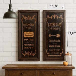 Puede incluir: Dos letreros negros enmarcados con letras y detalles dorados. Los letreros miden 30 cm de ancho y 70 cm de alto. Un letrero dice "Boulangerie A Paris" y el otro "Bistro Cafe A Paris". Los letreros están colgados en una pared de ladrillos blancos.