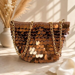 May include: A bronze-colored handbag covered in overlapping circular metallic sequins. The bag features a textured, crocheted base structure and two gold-toned chain straps that are attached to the top edges of the bag for carrying.