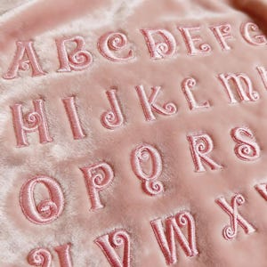 May include: Close-up of a pink, plush fabric embroidered with the alphabet in a decorative, cursive font. The letters are in a matching pink thread, creating a tone-on-tone effect. The fabric has a soft, velvety texture.