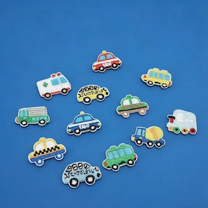 以下が含まれることがあります： 車、タクシー、救急車、スクールバス、電車など、さまざまな乗り物をモチーフにした刺繍ワッペン。赤、黄、緑、青などの鮮やかな色使いで、白と黒のディテールが施されています。青色の背景。
