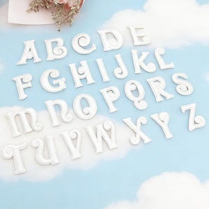May include: White alphabet letters, A to Z, with a decorative swirl design. The letters are displayed on a light blue background with a cloud pattern. The letters are made of a solid material.