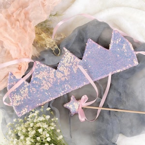 Può includere: Una corona di paillettes lavanda con nastri rosa e una bacchetta a forma di stella. La corona ha un design a tre punte. La bacchetta a stella è rosa e bianca con un fiocco di nastro rosa. Su sfondo grigio e bianco.