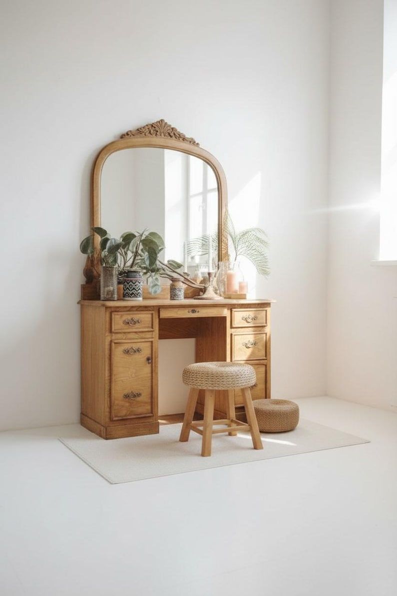 May include: A vintage wooden vanity with a large arched mirror, drawers, and a small stool. The vanity is made of light-colored wood and is decorated with plants and candles. The stool is made of woven material and wood.