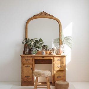 May include: A wooden vanity with an arched mirror, drawers, and a stool. The vanity is made of light-colored wood and has a decorative carved top. The stool is made of woven material and wood. Plants and candles are on the vanity.