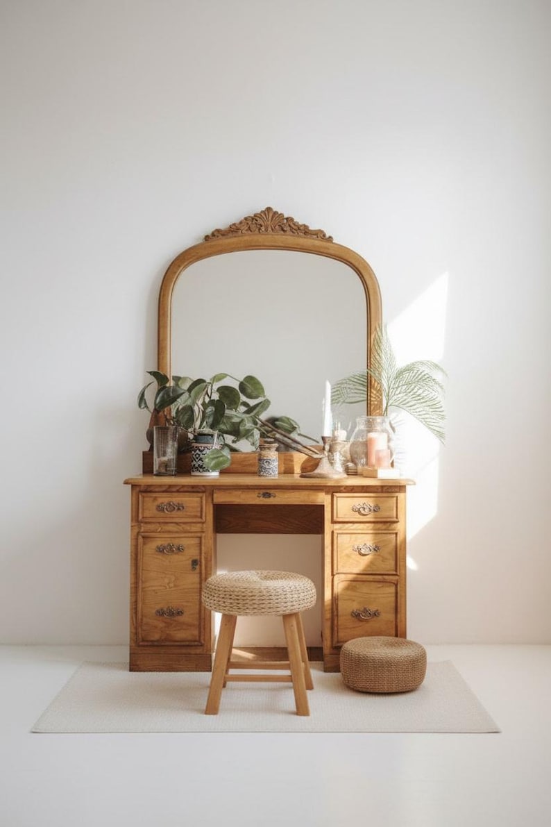 May include: A wooden vanity with an arched mirror, drawers, and a stool. The vanity is made of light-colored wood and has a decorative carved top. The stool is made of woven material and wood. Plants and candles are on the vanity.