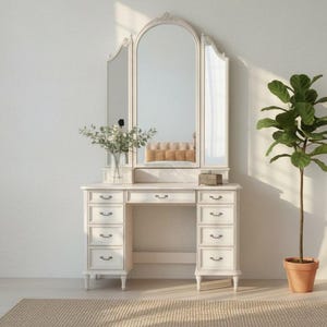 Puede incluir: Tocador blanco con espejo triple, cajones y una pequeña zona de mesa. Un jarrón con flores está sobre el tocador y una planta en maceta a la derecha. El tocador está sobre una alfombra beige.