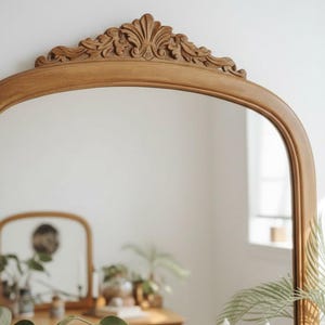 May include: A light brown, arched mirror with ornate carvings at the top. The mirror reflects a room with a wooden dresser, plants, and candles. The frame has a natural wood finish.