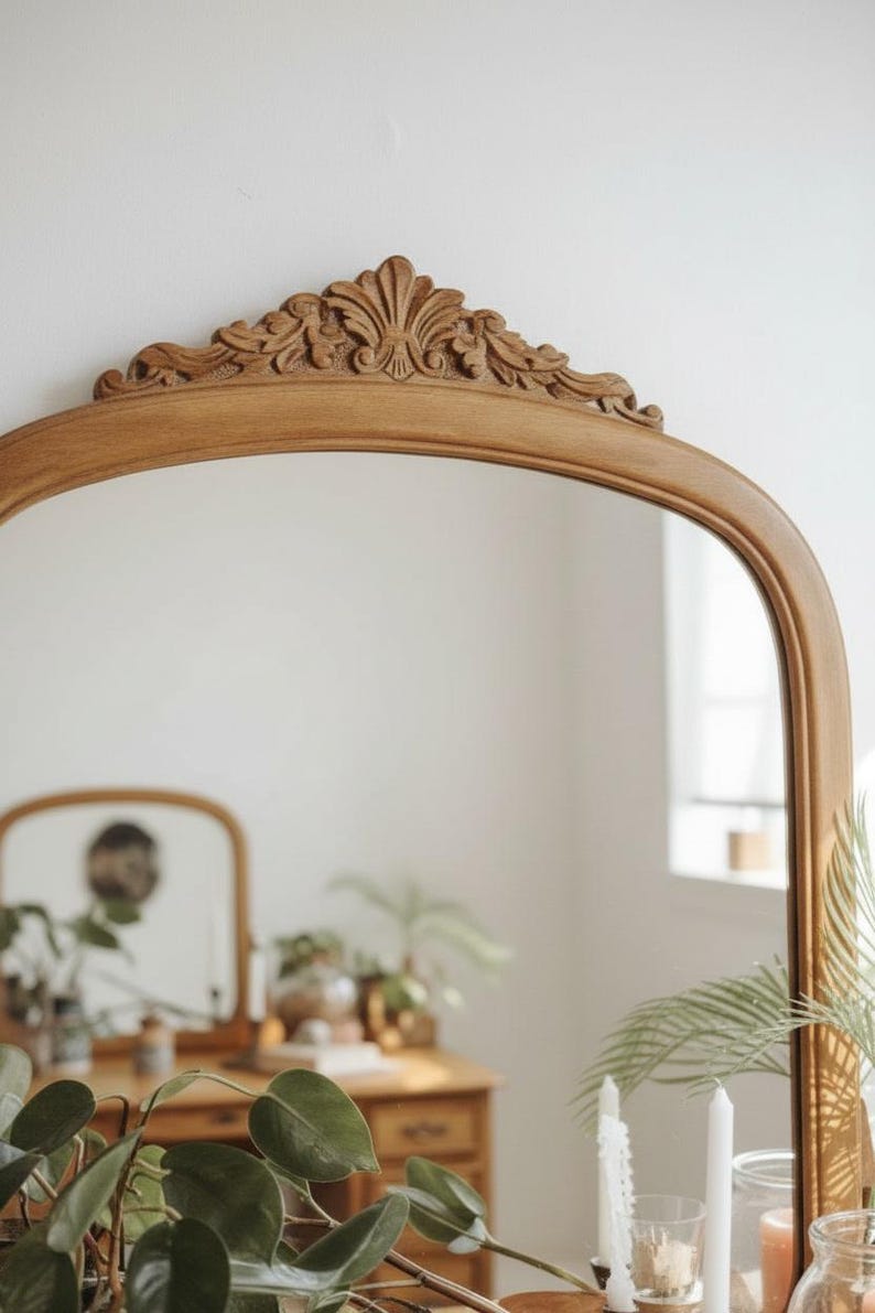 May include: A light brown, arched mirror with ornate carvings at the top. The mirror reflects a room with a wooden dresser, plants, and candles. The frame has a natural wood finish.