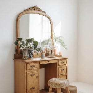 May include: A vintage wooden vanity with a large arched mirror, drawers, and a stool. The vanity is made of light-colored wood and has ornate detailing. The stool is woven with a natural fiber. Decorative items, including plants and candles, are on the vanity.