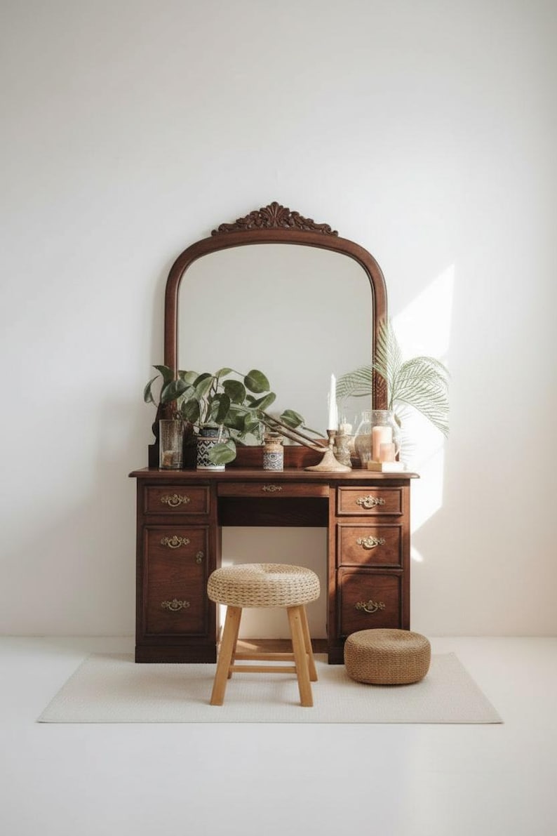 May include: A vintage wooden vanity with a large arched mirror, drawers, and decorative hardware. The vanity is adorned with plants, candles, and a woven stool. The overall aesthetic is warm and inviting, perfect for a bedroom or dressing area.