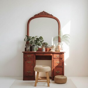 May include: A vintage wooden vanity with a large arched mirror, drawers, and a small stool. The vanity is a warm brown color, and the mirror has an ornate carved top. The stool is made of woven material and wood. Decorative plants and candles are on the vanity.