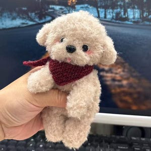 Peut inclure: Un jouet en peluche en forme de chien, de couleur crème, avec une écharpe bordeaux. Le jouet a une texture duveteuse, des yeux noirs et des joues roses. Le chien est tenu dans une main, avec un clavier visible en arrière-plan. Un jouet fait main.