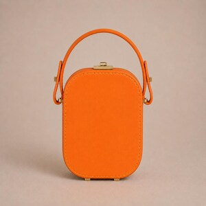 May include: A bright orange, vertically oriented, rectangular handbag with rounded corners. It features a single top handle attached with gold-toned metal hardware, a matching gold-toned clasp closure at the top, and visible stitching along the edges.