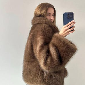 May include: A brown faux fur coat with wide sleeves and a large collar. The coat has a soft, textured appearance and a warm, neutral color. The coat is knee-length and appears to be made of a plush material.