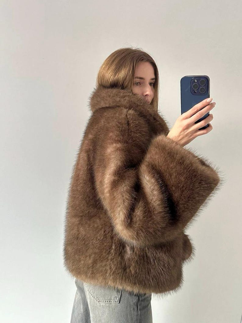 May include: A brown faux fur coat with wide sleeves and a large collar. The coat has a soft, textured appearance and a warm, neutral color. The coat is knee-length and appears to be made of a plush material.